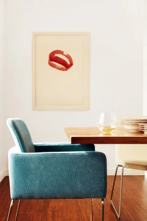 Interior view of a mid-century modern home with a blue chair, hardwood floor, and a framed lips art print on a white wall.
