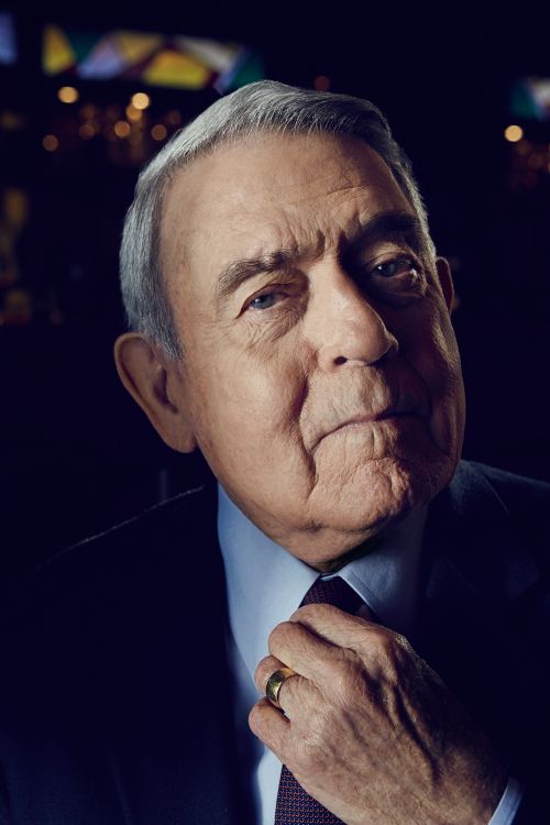 Headshot of an elderly gentleman with gray hair, wearing a suit and tie, and adjusting his tie knot. Dark background.