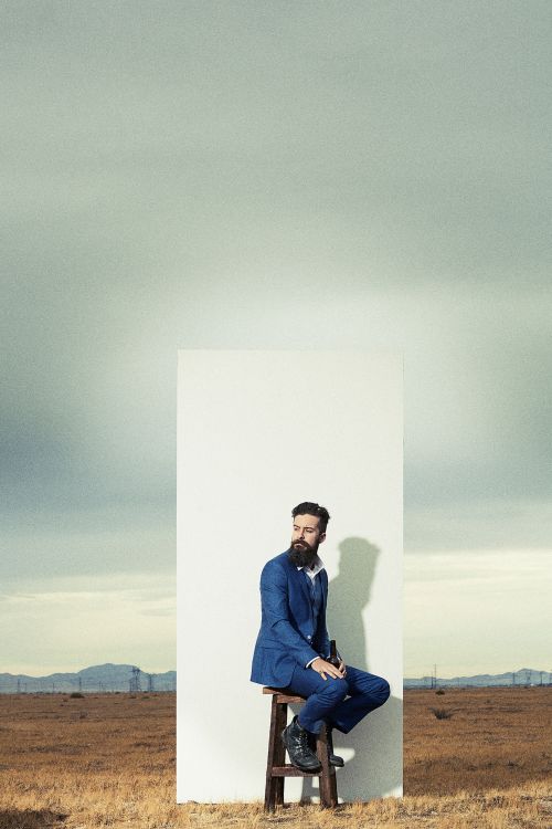 Man in a blue suit poses against a white backdrop in a dry, grassy field under a cloudy sky, seated on a wooden stool.