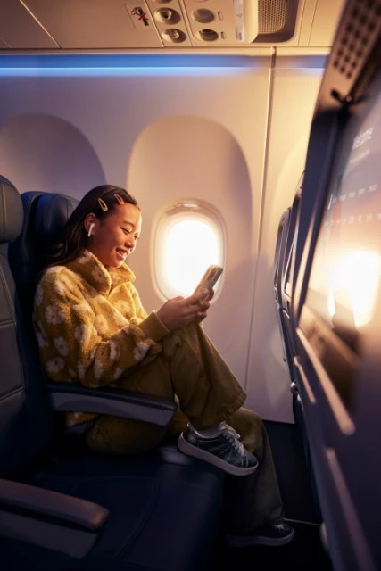 Young woman smiles while using a smartphone on an airplane, wearing headphones, with window and seats visible in the background.