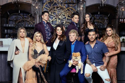 Group of nine adults and a dog posing indoors in formal attire with decorative gears and chandelier in the background.