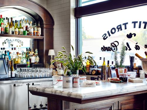 Brightly lit bar and restaurant interior features an archway with liquor bottles and stemware, a white marble countertop, and greenery.