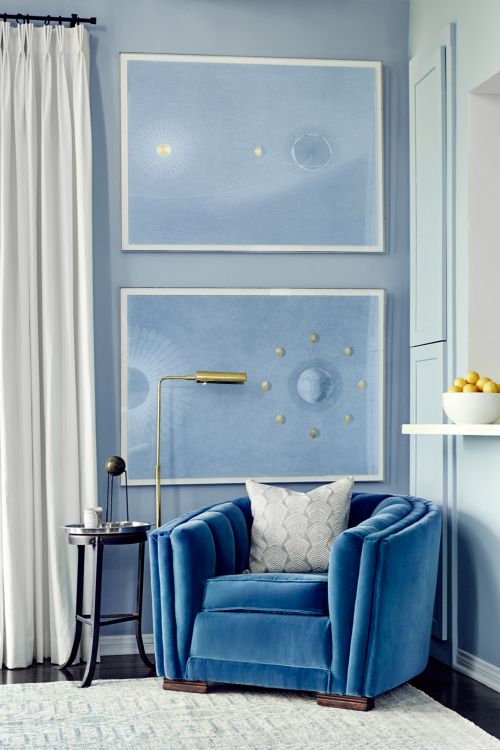 Blue velvet arm chair with accent table and brass floor lamp in a modern living room with blue walls and abstract art.