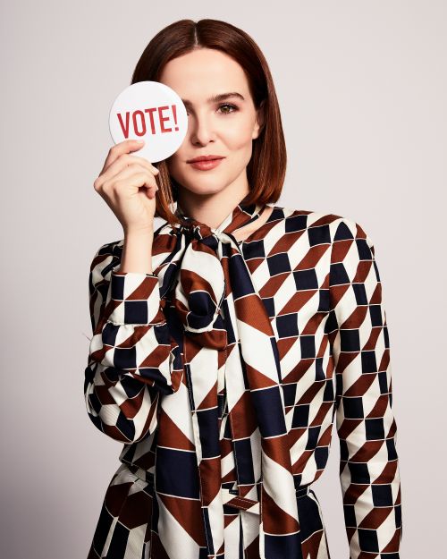 Headshot of fair-skinned woman holding a white circle sign that says Vote!, wearing a patterned blouse with a tie collar on a gray background.
