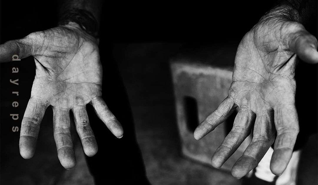 A black-and-white image featuring a close-up of two weathered human hands extended towards the viewer against a dark background.
