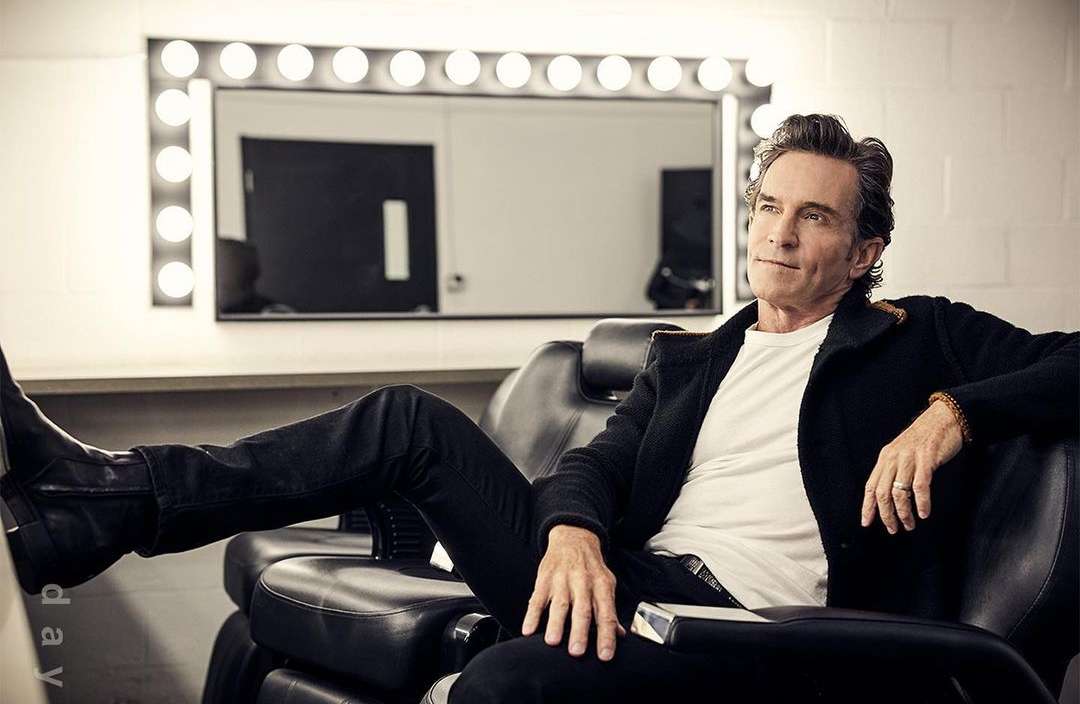 Adult male seated in a black chair with a white shirt and black coat. He is in a backstage area with a lighted mirror.