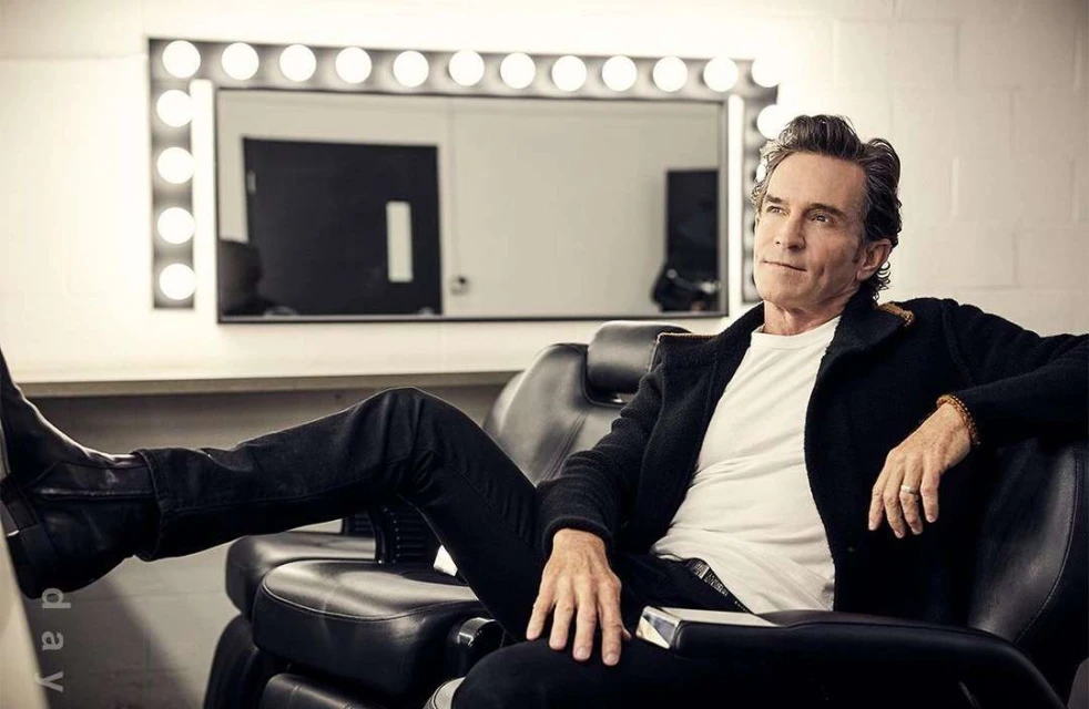 Adult male seated in a black chair with a white shirt and black coat. He is in a backstage area with a lighted mirror.
