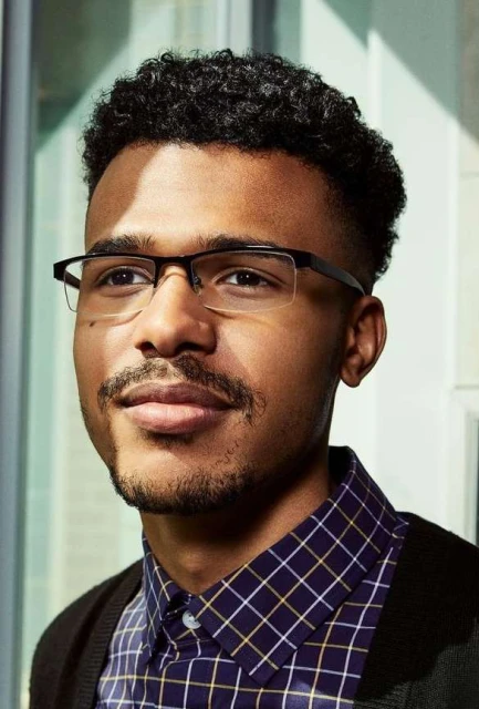 Close-up portrait of a Black adult male wearing glasses and a patterned shirt, looking off-camera with a neutral expression, indoors.