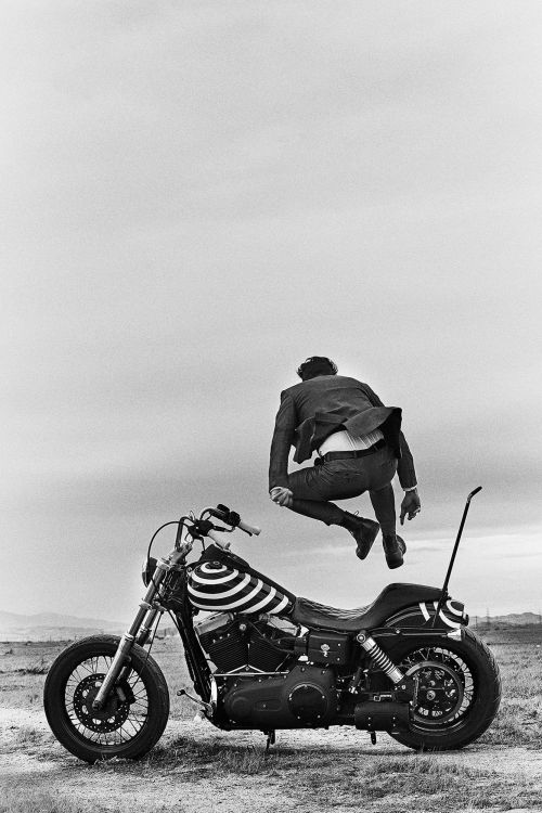 Black and white shot of a person leaping over a motorcycle in an outdoor desert landscape under a cloudy sky.