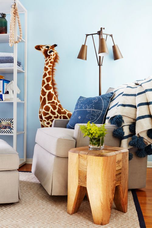 Interior of nursery with tan armchair, wooden side table with flowers, tall stuffed giraffe, blue throw pillow, and soft blue wall.