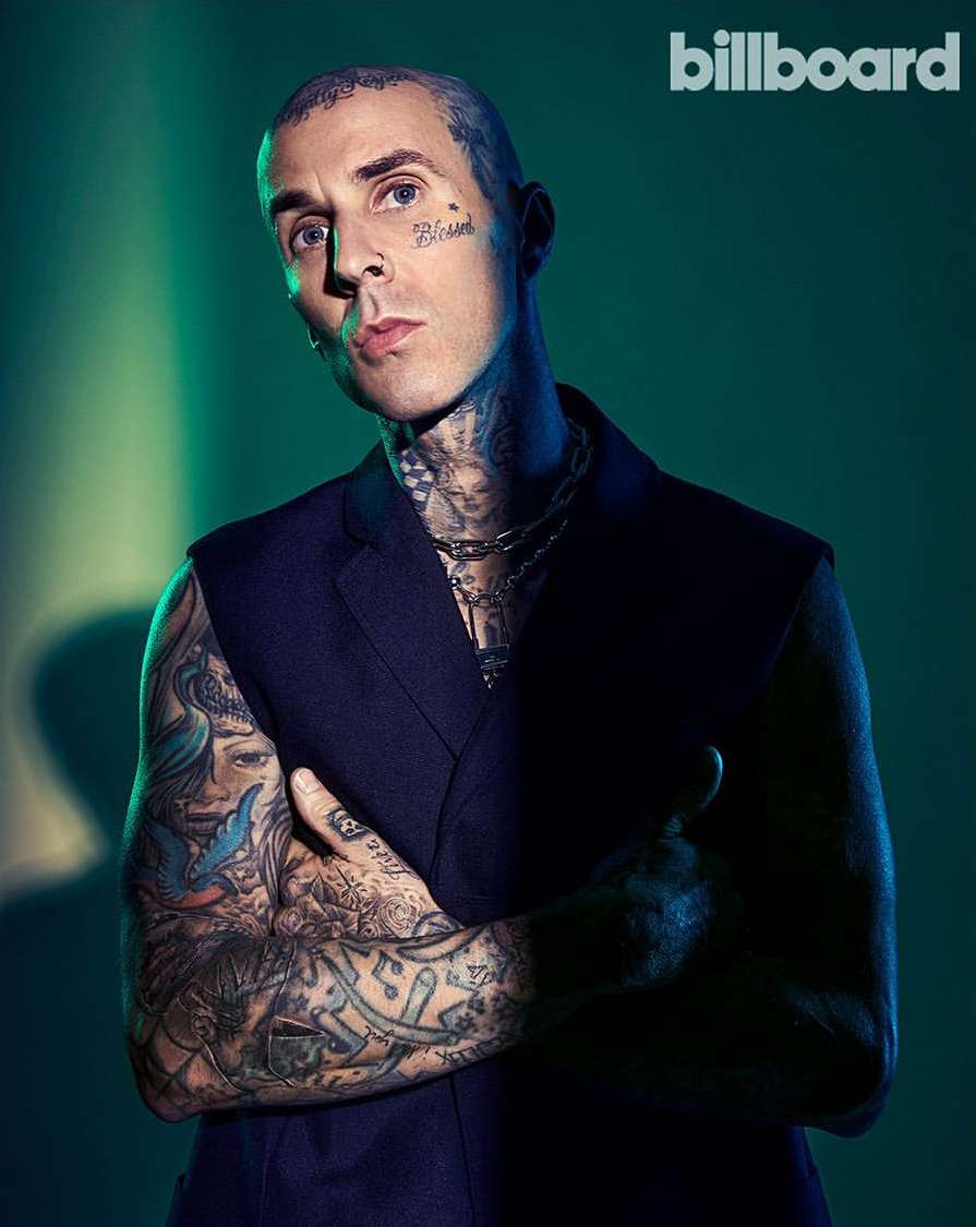 Portrait of tattooed adult male wearing a dark vest and silver chains with a serious expression against a teal-green background.