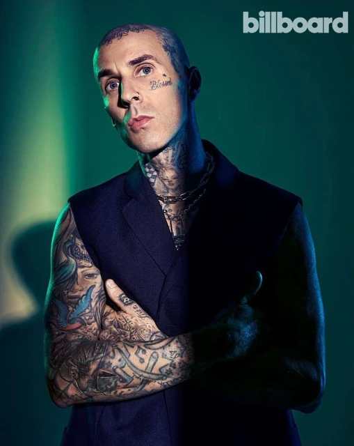 Portrait of tattooed adult male wearing a dark vest and silver chains with a serious expression against a teal-green background.