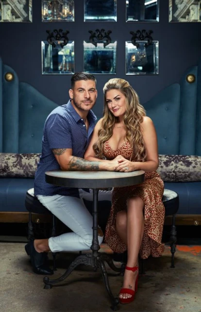 Portrait of a couple sitting at a table in a dimly lit interior setting. The man is wearing a button-down shirt and white pants. The woman is wearing a dress with a leopard print pattern.
