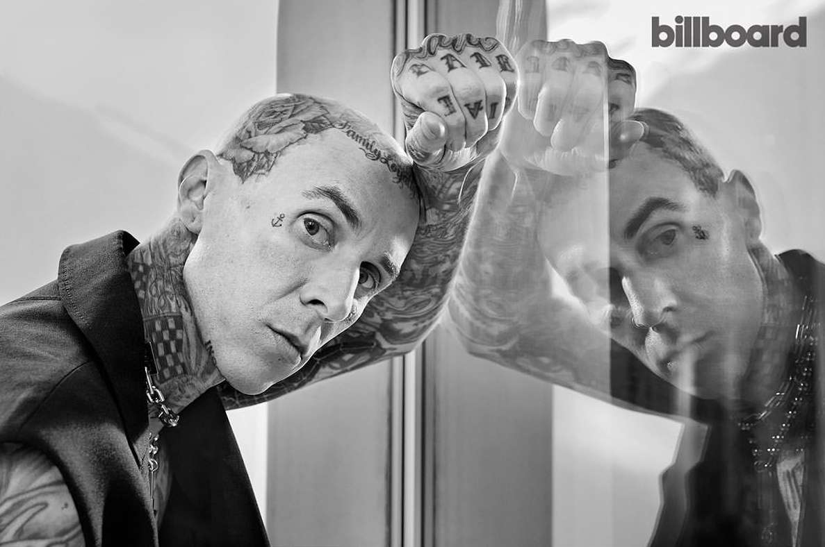 Black and white studio portrait of heavily tattooed adult male wearing a blazer, posing against glass with reflection, Billboard logo.