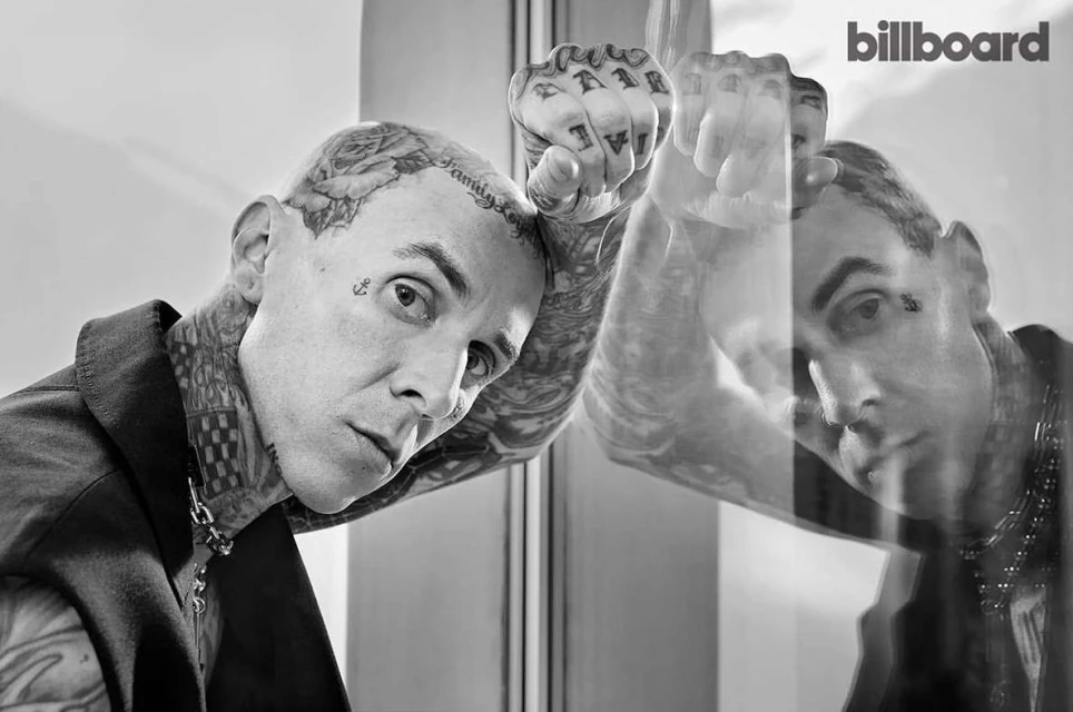 Black and white studio portrait of heavily tattooed adult male wearing a blazer, posing against glass with reflection, Billboard logo.
