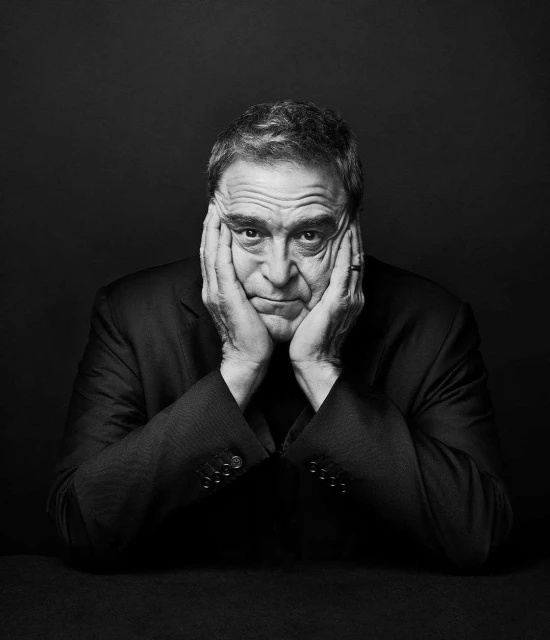 Black and white portrait of an adult male in a suit with his hands clasped to his face against a dark backdrop in a photo studio.