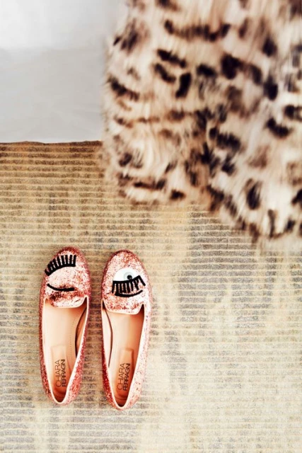 Close-up of a pair of pink glittery Chiara Ferragni slip-on shoes with eye design, on a striped rug near a fuzzy leopard-print throw.