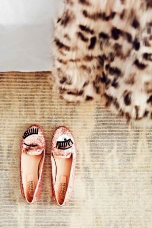 Close-up of a pair of pink glittery Chiara Ferragni slip-on shoes with eye design, on a striped rug near a fuzzy leopard-print throw.