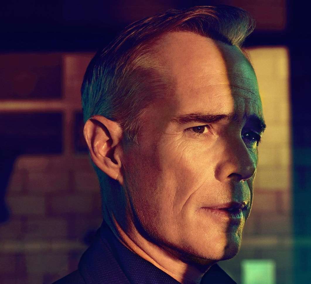 Headshot of a middle-aged man with fair skin in a dark suit, lit with yellow light on one side of his face in a dimly lit environment.