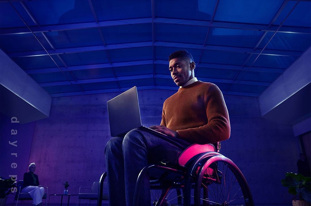Man in wheelchair working on laptop in modern, blue-lit interior with other people and decor in background. Focused and professional.