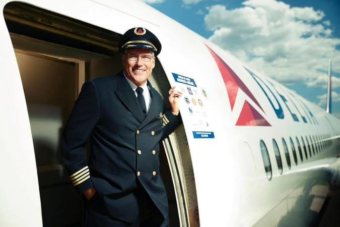 An airline pilot in uniform stands at the door of a Delta airplane, smiling and looking at the camera with a cloudy sky overhead.