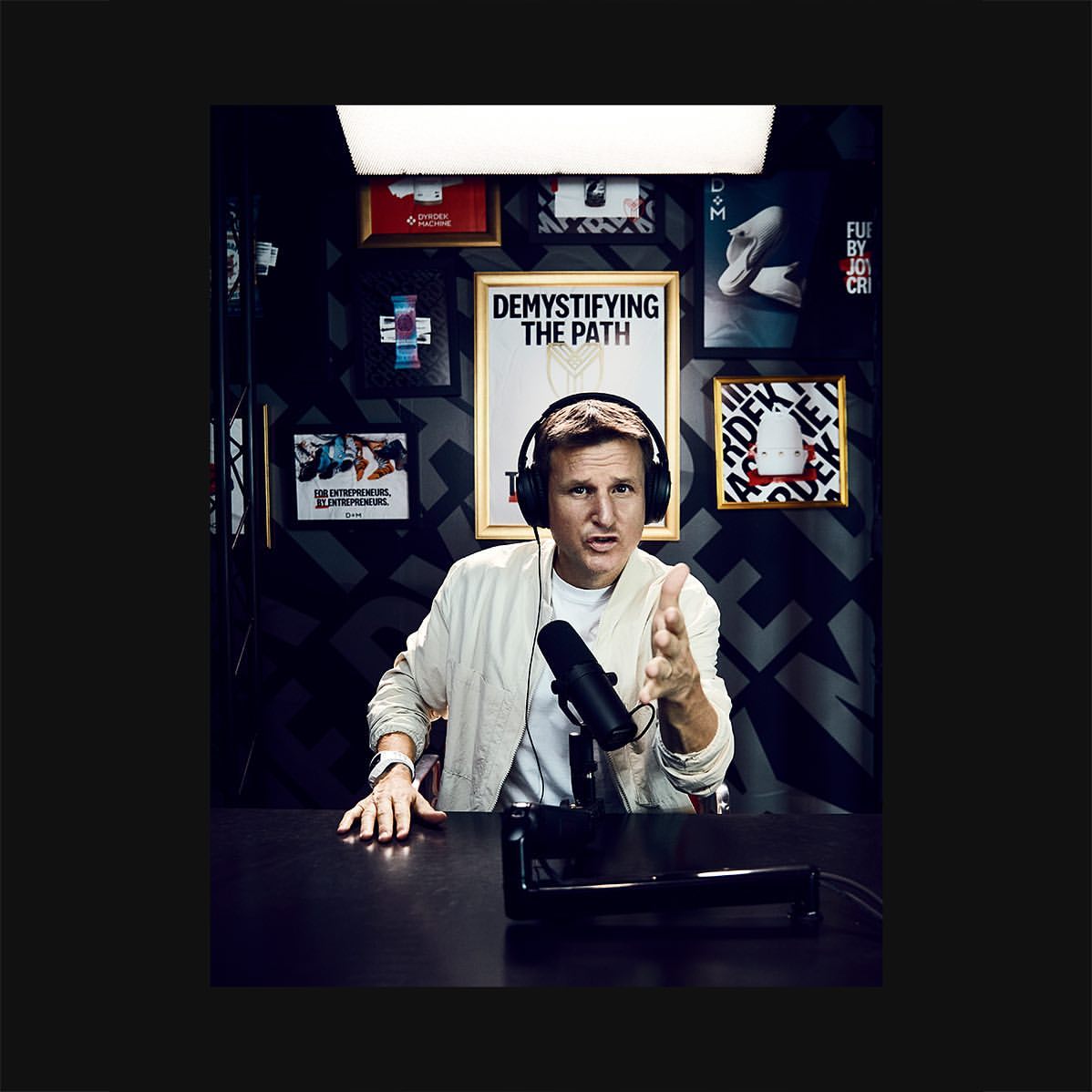 Adult male presenter with headphones speaks at a microphone in a podcast studio with framed posters and black patterned wallpaper.
