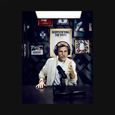 Adult male presenter with headphones speaks at a microphone in a podcast studio with framed posters and black patterned wallpaper.