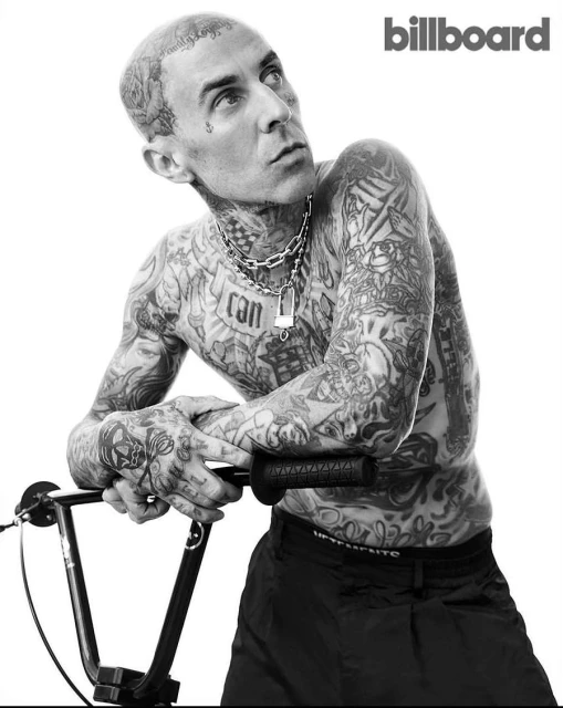 Black-and-white portrait of tattooed man resting arms on bicycle handlebars, wearing a chain necklace, posing in a photo studio.