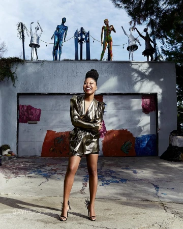 Black woman in a gold metallic dress standing outdoors smiling in front of a painted garage door with sculptures on top.