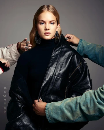 Portrait of a blonde woman wearing a black leather jacket with hands adjusting her collar against a grey background.