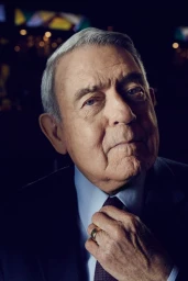 Close-up portrait of an elderly man adjusting his red patterned tie, wearing a dark suit jacket and light blue dress shirt