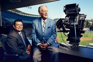 Two men in suits seated inside a broadcast booth overlooking an empty baseball stadium with a large television camera in the foreground.