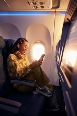 Young woman in a patterned yellow fuzzy jacket looking at her smartphone seated in an airplane cabin near a window