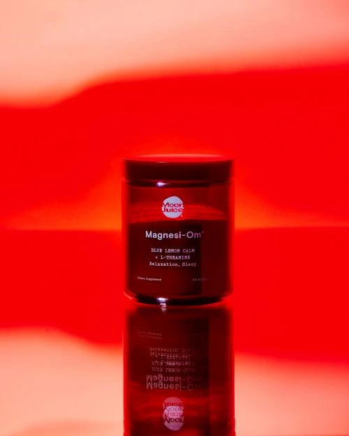 Moon Juice Magnesi-Om supplement jar on a reflective surface against a red and orange gradient background in a studio setting.