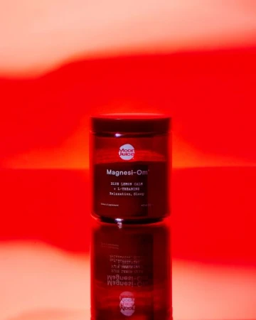 Moon Juice Magnesi-Om supplement jar on a reflective surface against a red and orange gradient background in a studio setting.