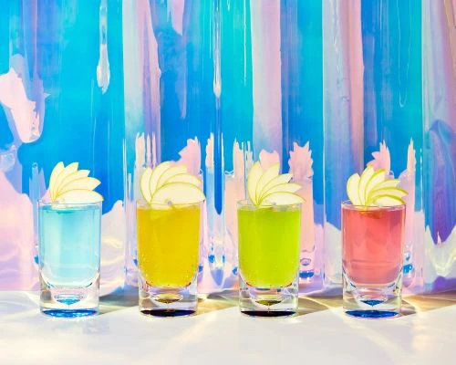 Colorful shot glasses filled with different liquids and garnished with sliced apples against an iridescent background creating a vibrant and eye-catching scene.