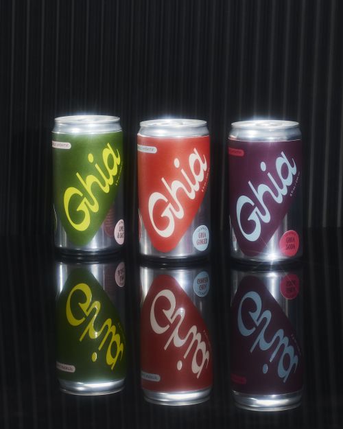 Three Ghia soda cans in green, red, and purple arranged on a reflective surface against a black background. Food and beverage still life.