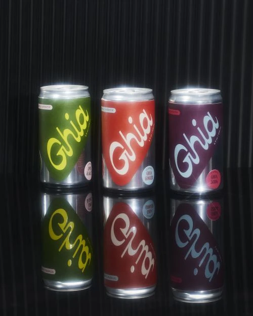 Three Ghia soda cans in green, red, and purple arranged on a reflective surface against a black background. Food and beverage still life.