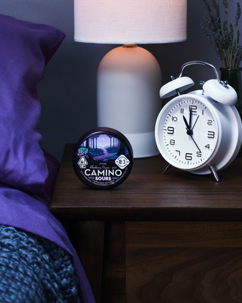 Still life of Camino Sours cannabis product on a wooden nightstand with a white alarm clock, lamp, and partially visible bed.