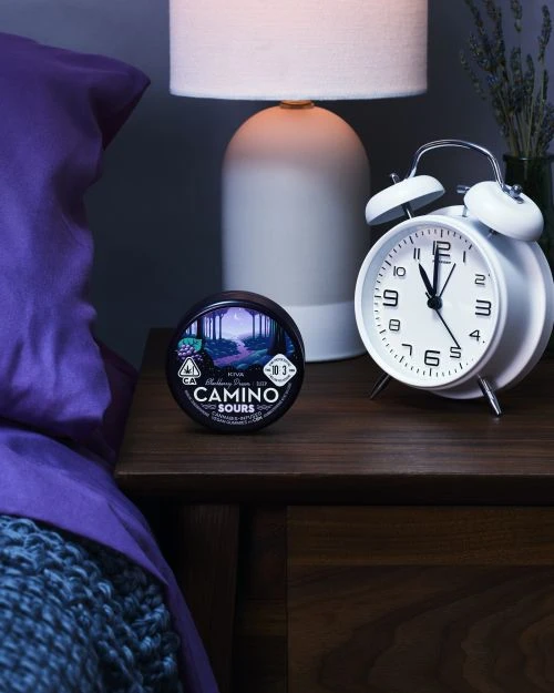 Still life of Camino Sours cannabis product on a wooden nightstand with a white alarm clock, lamp, and partially visible bed.