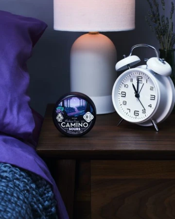 Still life of Camino Sours cannabis product on a wooden nightstand with a white alarm clock, lamp, and partially visible bed.