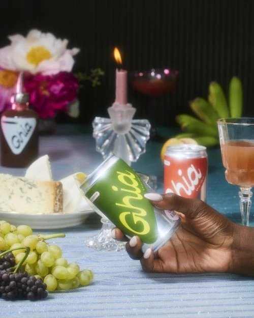 Hand holding a Ghia beverage can amid a table setting with fruit, cocktails, desserts, and a lit candle in an intimate setting.