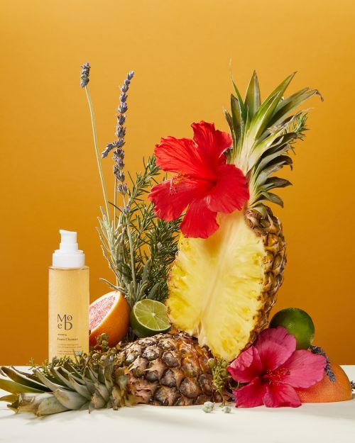Still life with Mōd Beauty product bottle, pineapple halves, grapefruit, hibiscus, lime, and lavender on a white surface against yellow backdrop.