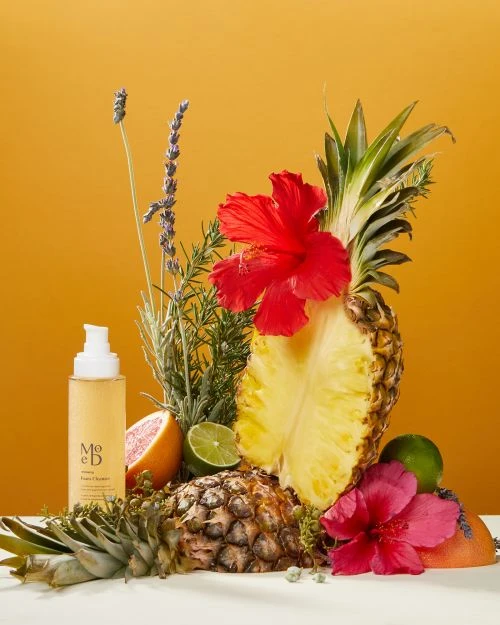 Still life with Mōd Beauty product bottle, pineapple halves, grapefruit, hibiscus, lime, and lavender on a white surface against yellow backdrop.