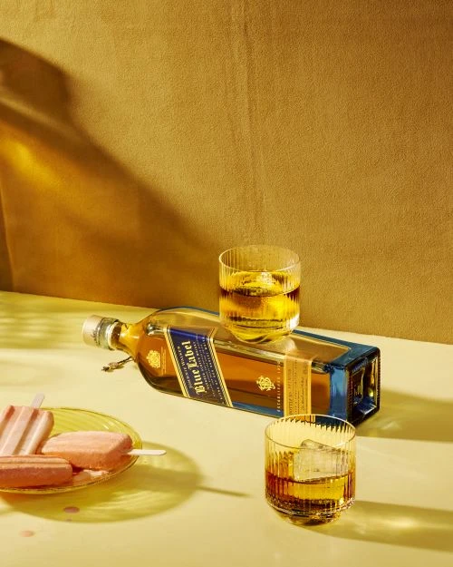 Still life featuring a bottle of Johnnie Walker Blue Label whisky, two glasses with ice, and popsicles on a yellow surface against a gold backdrop.
