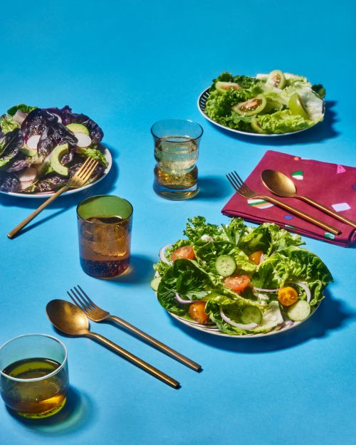 Overhead shot of vibrant salads and drinks on a bright blue table, featuring fresh greens, tomatoes, cucumbers, and golden cutlery.