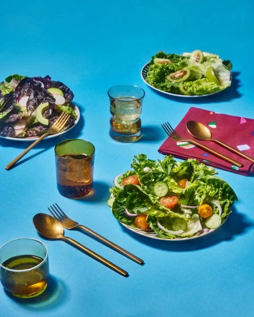Overhead shot of vibrant salads and drinks on a bright blue table, featuring fresh greens, tomatoes, cucumbers, and golden cutlery.