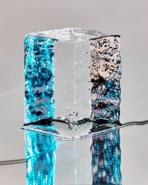 Abstract cube with textured surface, illuminated with blue and silver light, casting a reflection, against a light grey backdrop.