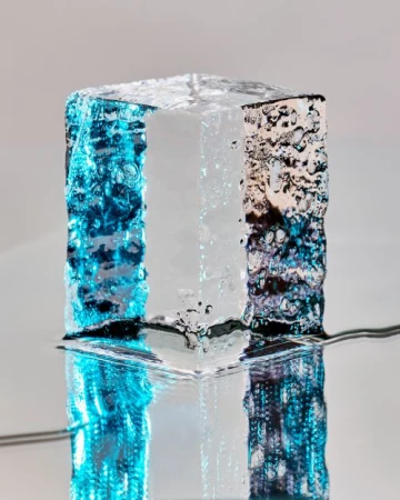 Abstract cube with textured surface, illuminated with blue and silver light, casting a reflection, against a light grey backdrop.