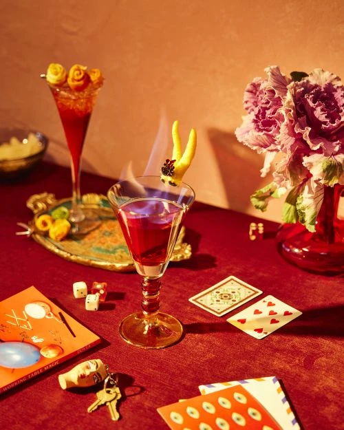 Two red cocktails on a table, one flambé, with dice, playing cards, flowers, and an orange book featuring vintage-style lettering.